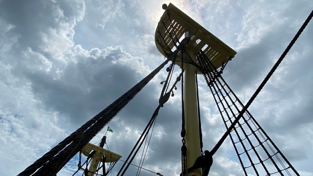 Skyward view of two light yellow ship masts with black rope attached and sun peaking through clouds.